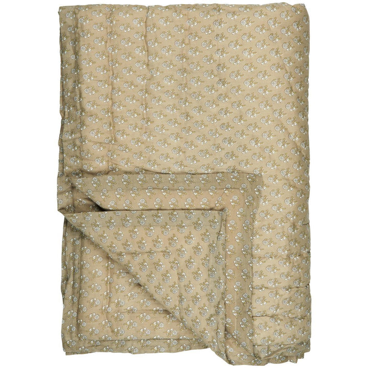 Une grande couverture beige ornée d'un motif floral discret, composée de petites fleurs blanches et vertes, est soigneusement étalée. Un coin de la couverture est replié pour montrer l'épaisseur du tissu et la texture matelassée, ajoutant une touche de confort visuel.