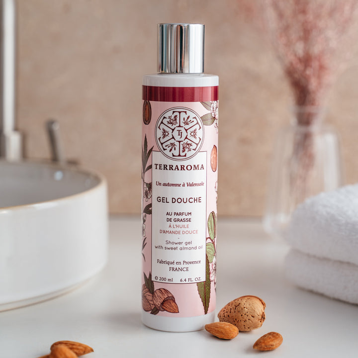 L'image montre un flacon de gel douche de la gamme "Un Automne à Valensole" de la marque Terraroma. Ce gel douche est parfumé au parfum de Grasse et enrichi en huile d'amande douce, réputée pour ses propriétés nourrissantes. Le flacon de 200 ml présente un design délicat avec des motifs d'amandes et des couleurs douces rappelant l'automne, dans des tons roses et marron. Autour du flacon, quelques amandes entières et en coque sont disposées, soulignant l'ingrédient clé du produit.