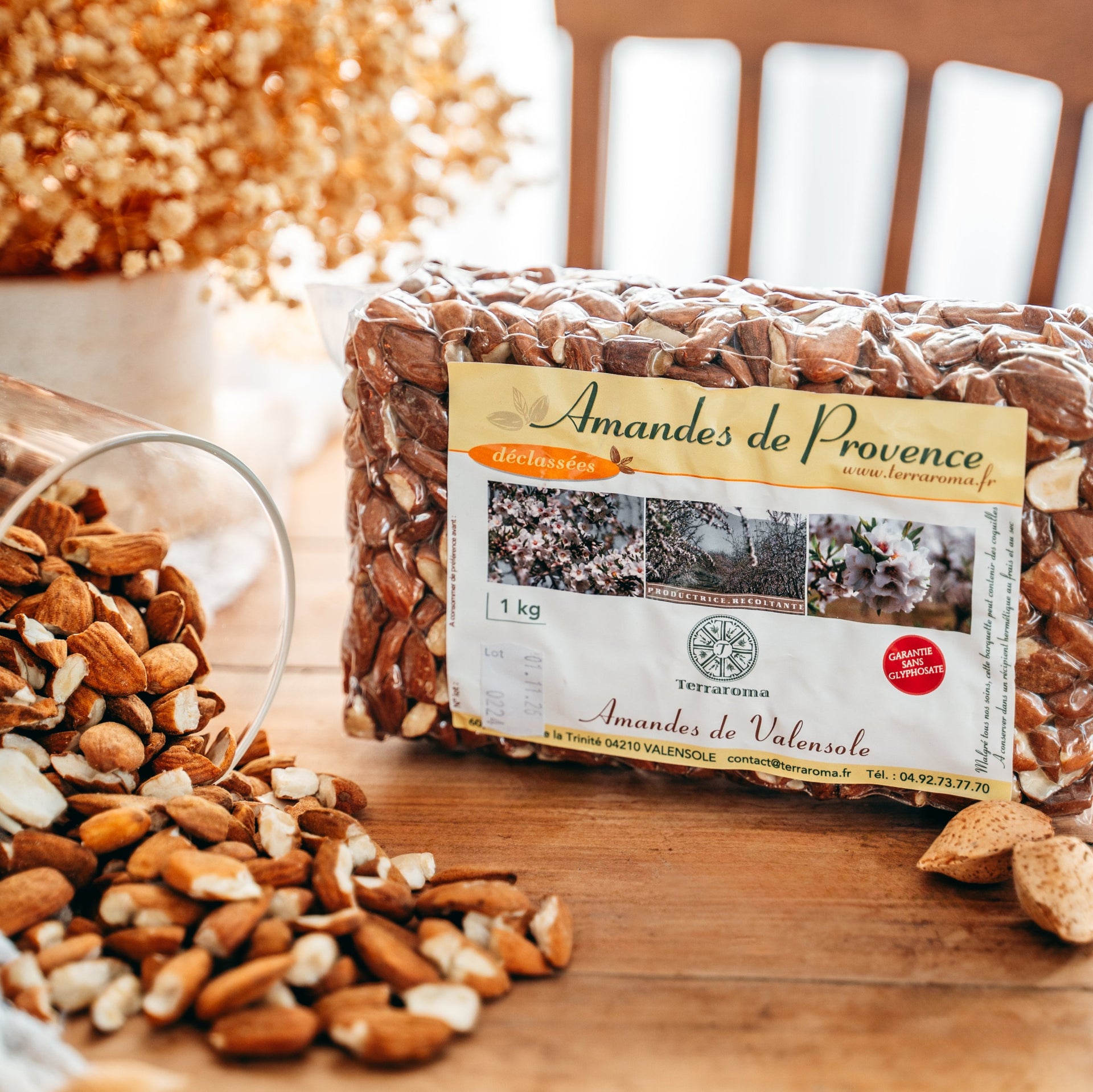 L'image montre un sachet de 1 kg d'amandes de Provence déclassées de la marque Terraroma, étiqueté "Amandes de Valensole". Le sachet est posé sur une table en bois, entouré d'amandes entières et éclatées, avec un verre renversé. L'arrière-plan est décoré de fleurs séchées et baigné de lumière naturelle.