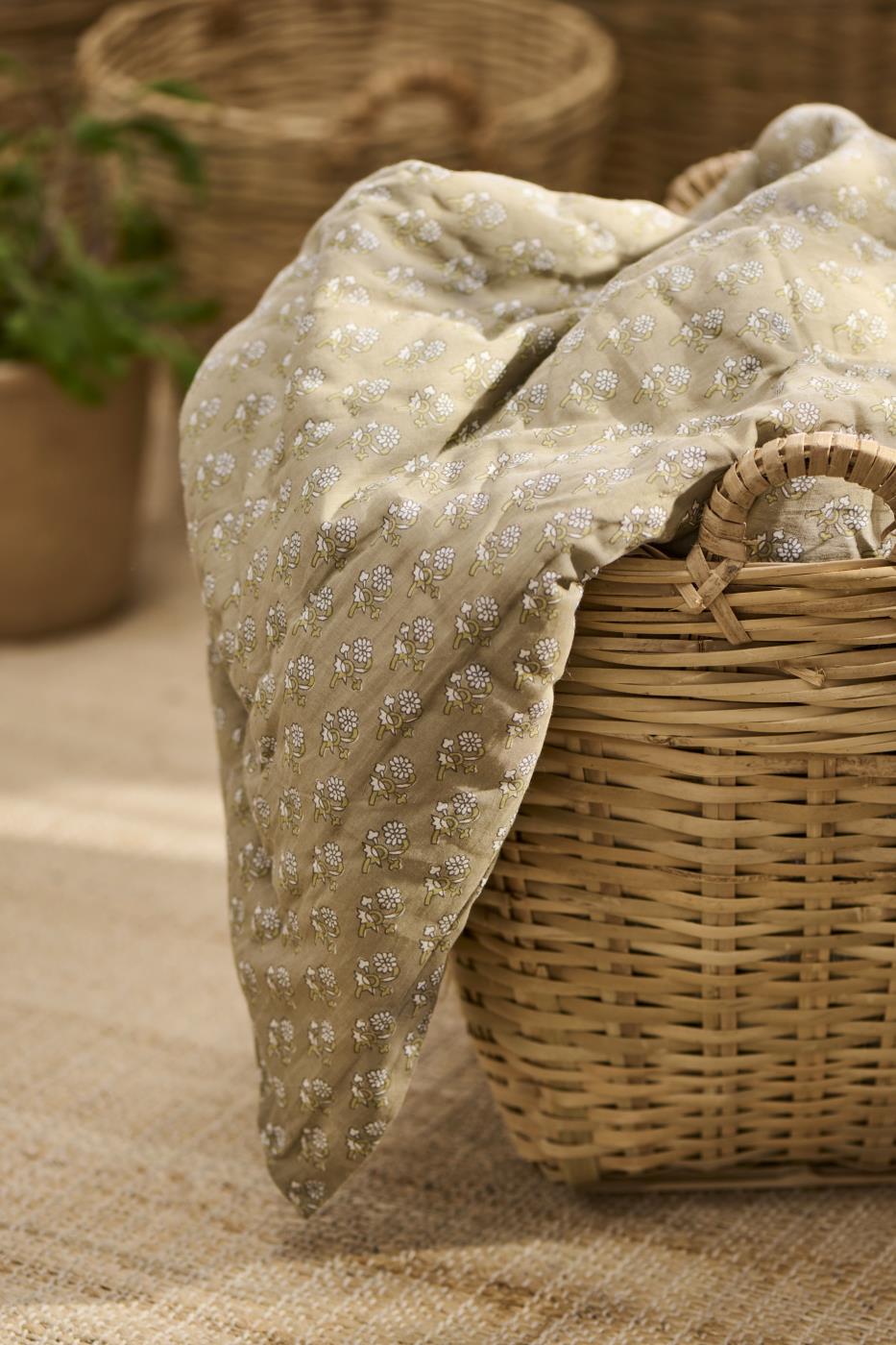 Un panier en osier tressé contient une couverture en tissu beige avec un motif floral blanc et vert. La couverture est légèrement drapée sur le bord du panier, exposant ses détails délicats. En arrière-plan, d'autres paniers en osier sont visibles, contribuant à l'atmosphère naturelle et chaleureuse de la scène.