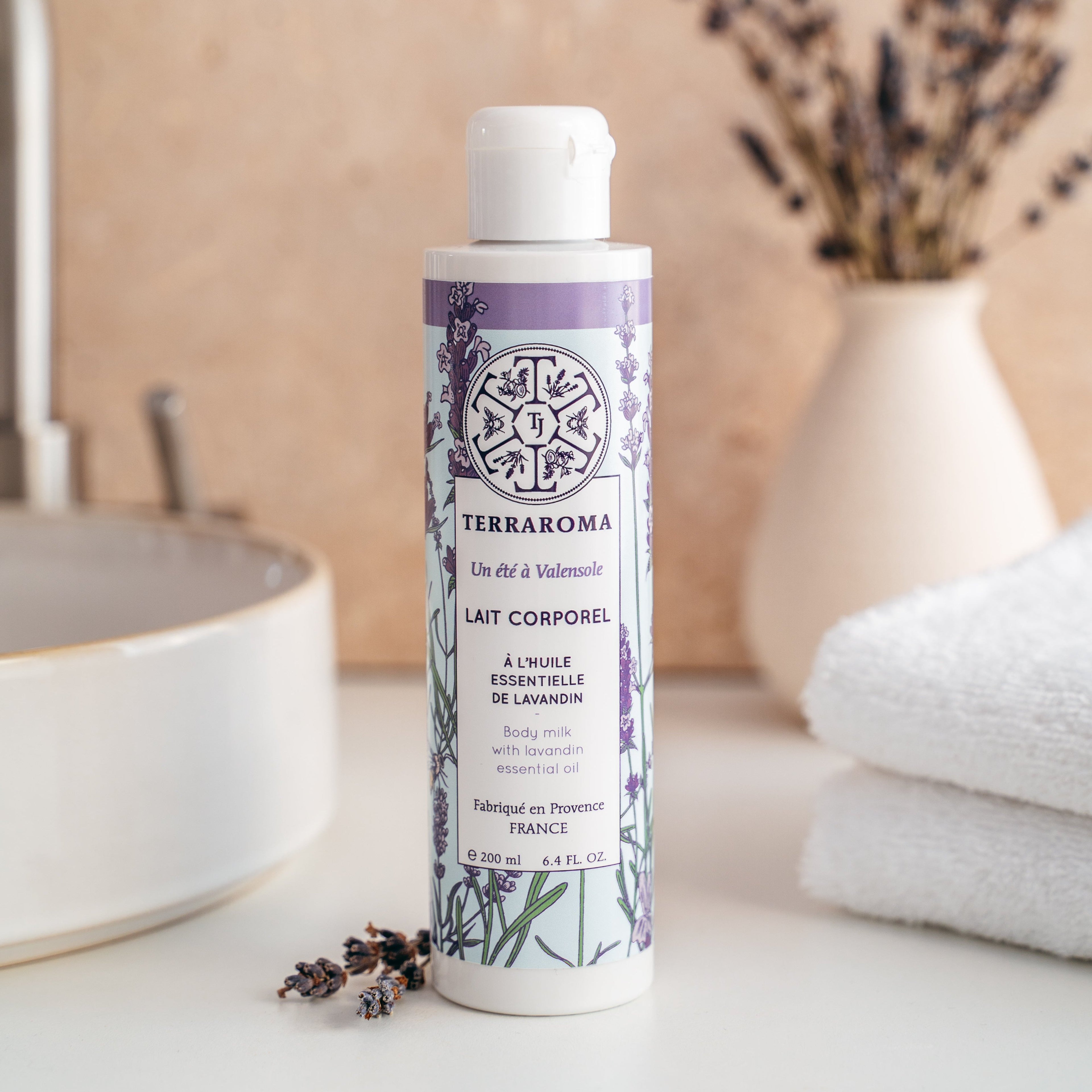 L'image montre un flacon de lait corporel de la gamme "Un Été à Valensole" de la marque Terraroma. Ce produit est formulé à l'huile essentielle de lavandin, connue pour ses propriétés apaisantes et hydratantes. Le flacon de 200 ml présente un design élégant avec des motifs de brins de lavande sur un fond violet, rappelant l'origine provençale du produit. Quelques brins de lavande séchée sont disposés à côté du flacon, renforçant l'atmosphère naturelle et apaisante. En arrière-plan, un évier et une serviette