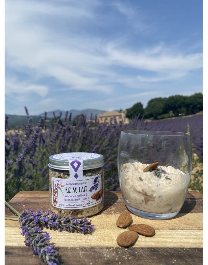 Sur cette image, on voit un pot en verre de préparation pour riz au lait aux amandes grillées et à la lavande de Provence posé sur une planche en bois. À côté du pot, un verre transparent contient du riz au lait crémeux, décoré d'une amande et de fleurs de lavande, ajoutant une touche visuelle provençale et raffinée. En arrière-plan, un champ de lavande s'étend sous un ciel bleu partiellement nuageux, avec une maison provençale au loin. 