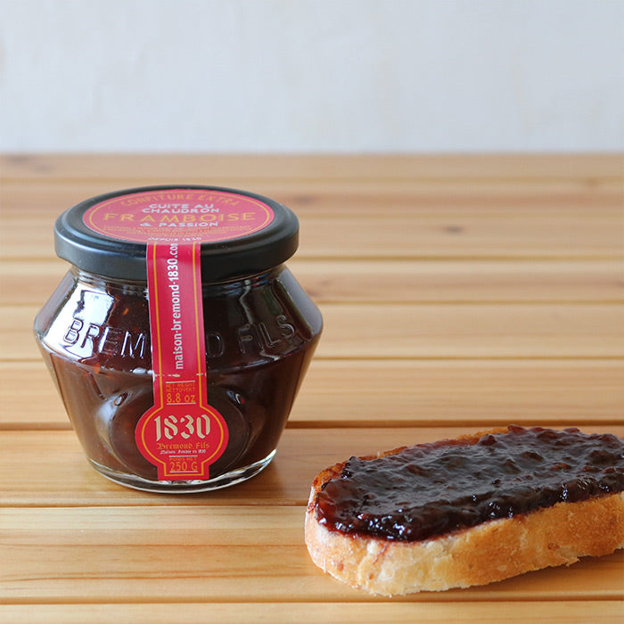 Un pot en verre contenant de la confiture extra de framboise et passion de la marque Maison Brémond, avec un couvercle noir et une étiquette rouge indiquant 'Confiture extra framboise & passion' et un poids net de 250 g. À côté du pot, une tranche de pain est tartinée de cette confiture, montrant sa texture lisse et brillante. Le fond est en bois, créant une ambiance chaleureuse et naturelle.