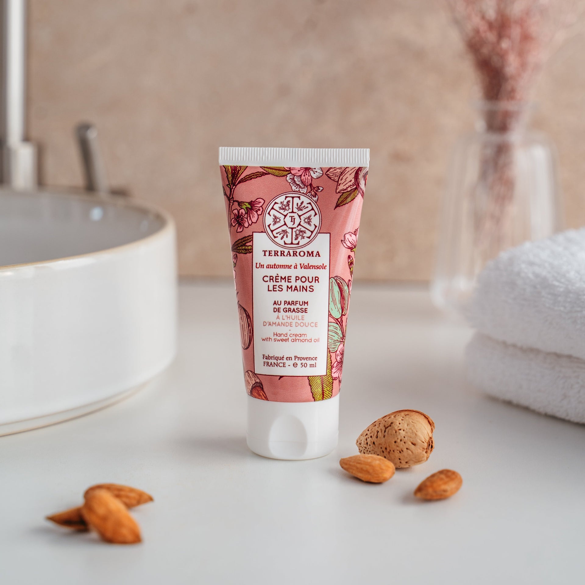 L'image montre un tube de crème pour les mains de la gamme "Un Automne à Valensole" de la marque Terraroma. Cette crème est parfumée au parfum de Grasse et enrichie en huile d'amande douce, reconnue pour ses propriétés hydratantes et apaisantes. Le tube de 50 ml arbore un design floral dans des tons roses et doux, évoquant la saison automnale et la douceur de l'amande. Autour du tube, quelques amandes et coquilles d'amande sont disposées, renforçant l'idée d'un produit naturel.