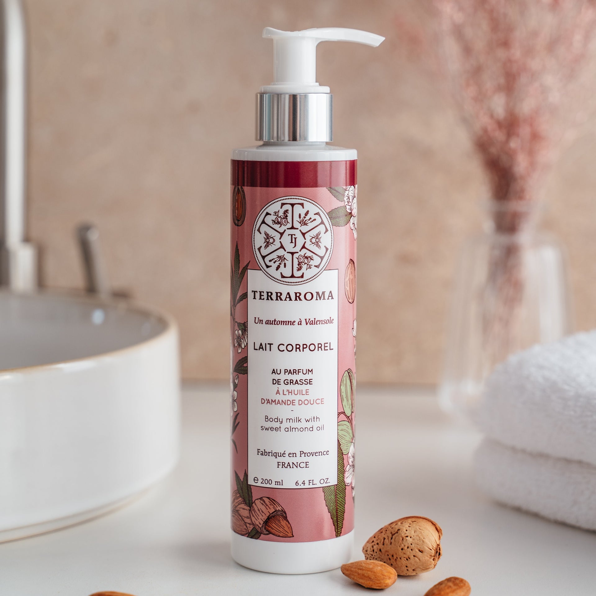 L'image montre un flacon de lait corporel de la gamme "Un Automne à Valensole" de la marque Terraroma. Ce lait corporel est parfumé au parfum de Grasse et enrichi en huile d'amande douce, connue pour ses propriétés hydratantes et adoucissantes. Le flacon de 200 ml, avec une pompe pratique, présente un design délicat aux motifs d'amandes sur un fond rose doux, reflétant l'essence automnale du produit. Autour du flacon, quelques amandes entières et en coque sont disposées, soulignant la naturalité.